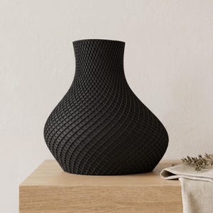 May include: A black vase with a unique, textured surface. The vase has a wide base that tapers to a narrow neck. The surface is covered in a repeating geometric pattern. The vase is sitting on a wooden surface.