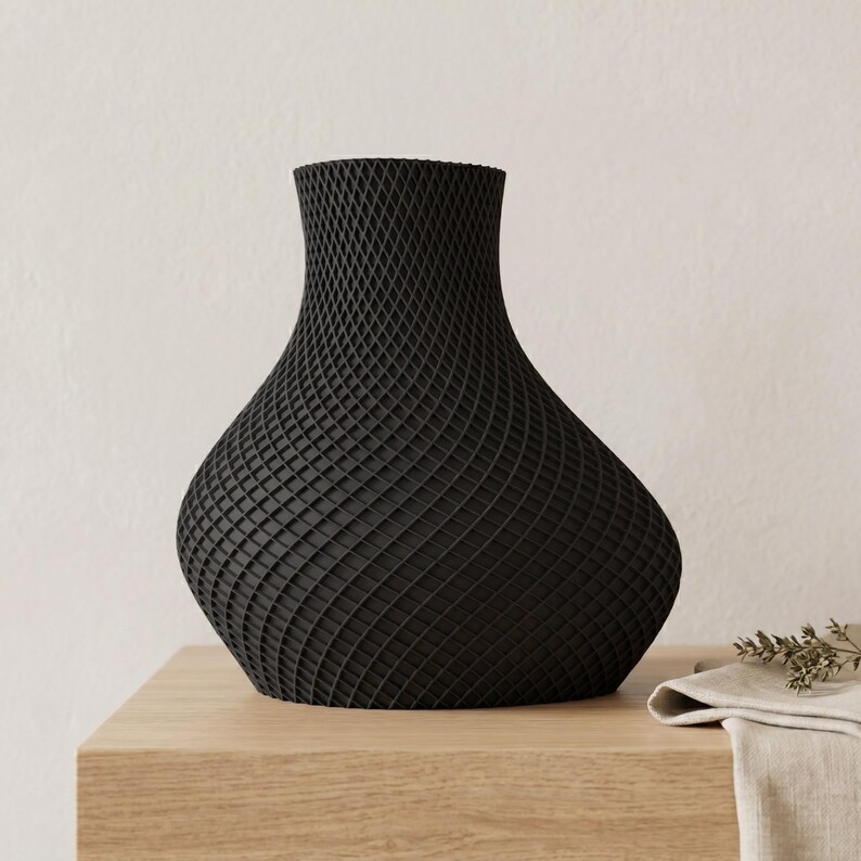 May include: A black vase with a unique, textured surface. The vase has a wide base that tapers to a narrow neck. The surface is covered in a repeating geometric pattern. The vase is sitting on a wooden surface.