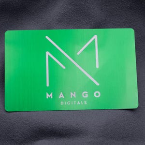Eine einzelne grüne Visitenkarte aus Aluminium mit lasergraviertem Logo "MANGO DIGITALS". Das minimalistische Design in Weiß kontrastiert mit der metallisch glänzenden Oberfläche. Die hochwertige Gravur verleiht der Karte ein elegantes Erscheinungsbild, ideal für moderne Marken und kreative Unternehmer, die Wert auf edles, langlebiges Design legen und einen bleibenden Eindruck hinterlassen möchten.
