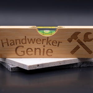 Op de afbeelding: Houten waterpas met de tekst "Handwerker Genie" en een hamer- en moersleutelsymbool. De waterpas heeft een bubbelindicator. Het ligt op twee stenen tegels.
