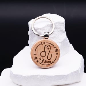 Ein gravierter Holz-Schlüsselanhänger mit dem Sternzeichen Löwe wird auf weißem Steinpodest präsentiert. Die runde Holzform zeigt das astrologische Symbol mit Sternen, Mond und Schriftzug „Löwe“. Ein stabiler Schlüsselring aus Metall ist oben befestigt. Das Bild betont die detailreiche Gravur, die warme Holzstruktur und die hochwertige Verarbeitung, wodurch der Schlüsselanhänger zu einem einzigartigen, personalisierten Geschenk wird.