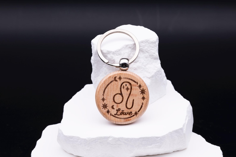 Ein gravierter Holz-Schlüsselanhänger mit dem Sternzeichen Löwe wird auf weißem Steinpodest präsentiert. Die runde Holzform zeigt das astrologische Symbol mit Sternen, Mond und Schriftzug „Löwe“. Ein stabiler Schlüsselring aus Metall ist oben befestigt. Das Bild betont die detailreiche Gravur, die warme Holzstruktur und die hochwertige Verarbeitung, wodurch der Schlüsselanhänger zu einem einzigartigen, personalisierten Geschenk wird.