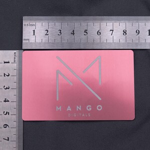 Roségoldene Metallkarte mit Mango-Logo, neben Linealen zur Größenangabe fotografiert. Die Maße entsprechen klassischen Kreditkarten (8,6 × 5,4 cm). Robuste, langlebige und moderne Alternative zu Papierkarten, ideal als auffällige Businesskarte oder kreative Kundendankeskarte im exklusiven Design.