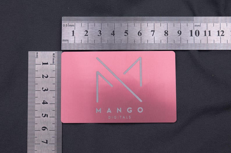 Roségoldene Metallkarte mit Mango-Logo, neben Linealen zur Größenangabe fotografiert. Die Maße entsprechen klassischen Kreditkarten (8,6 × 5,4 cm). Robuste, langlebige und moderne Alternative zu Papierkarten, ideal als auffällige Businesskarte oder kreative Kundendankeskarte im exklusiven Design.
