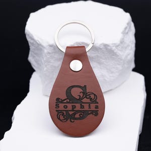 Personalisierter Schlüsselanhänger aus hochwertigem braunem Kunstleder mit individueller Lasergravur. Das elegante Monogramm mit Namen wie „Sophia“ verleiht dem Accessoire eine edle, persönliche Note. Durch den robusten Metallring eignet sich der Anhänger ideal für Auto-, Haus- oder Büroschlüssel. Perfekt als Geschenk für Männer, Frauen oder Paare, die stilvolle und langlebige Accessoires schätzen.