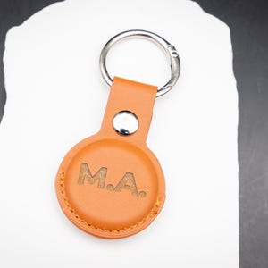 Brauner AirTag Schlüsselanhänger aus PU Leder mit individueller Lasergravur der Initialen M A. Der Schlüsselanhänger liegt auf einem weißen Stein und zeigt die hochwertige Lederstruktur sowie saubere Nähte. Perfekt geeignet für Apple AirTag und ideal als personalisiertes Geschenk für Männer Frauen oder Paare.