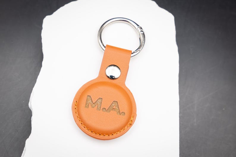Brauner AirTag Schlüsselanhänger aus PU Leder mit individueller Lasergravur der Initialen M A. Der Schlüsselanhänger liegt auf einem weißen Stein und zeigt die hochwertige Lederstruktur sowie saubere Nähte. Perfekt geeignet für Apple AirTag und ideal als personalisiertes Geschenk für Männer Frauen oder Paare.