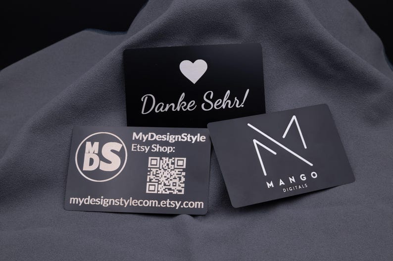 Schwarze Visitenkarten aus Aluminium-Metall mit matter Oberfläche, lasergraviert mit Name, Logo, Grafik oder QR-Code. Die Karten im Kreditkartenformat (ca. 8,6 × 5,4 cm) sind robust, leicht und langlebig. Die Darstellung zeigt mehrere Karten, u. a. mit „Danke Sehr!“-Schriftzug, Firmenlogo und QR-Code auf edlem Stoffhintergrund. Ideal als moderne Business- und Networking-Karten mit hochwertigem Look.