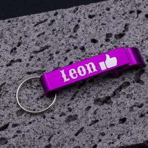 ila Flaschenöffner Schlüsselanhänger aus Aluminium mit Gravur „Leon“ und Daumen-hoch-Symbol. Praktischer Kapselheber mit Schlüsselring, stabil und leicht, perfekt als personalisiertes Geschenk für Freunde oder Familie. Individuell gravierbar mit Namen, Symbolen oder Logos, modernes Accessoire für Schlüsselbund und Flaschenöffnen, dargestellt auf grauem Steinuntergrund.