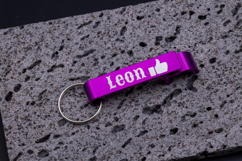 ila Flaschenöffner Schlüsselanhänger aus Aluminium mit Gravur „Leon“ und Daumen-hoch-Symbol. Praktischer Kapselheber mit Schlüsselring, stabil und leicht, perfekt als personalisiertes Geschenk für Freunde oder Familie. Individuell gravierbar mit Namen, Symbolen oder Logos, modernes Accessoire für Schlüsselbund und Flaschenöffnen, dargestellt auf grauem Steinuntergrund.