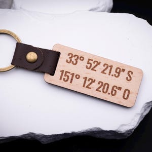 in gravierter Holz Schlüsselanhänger mit den Koordinaten „33° 52' 21.9'' S, 151° 12' 20.6'' O“ liegt auf weißem Stein. Der Anhänger ist aus hellem Holz gefertigt, mit braunem Lederband und stabilem Metallring in Antik-Gold versehen. Perfekt für personalisierte Gravuren von besonderen Orten, Namen oder Sprüchen. Ein liebevolles Geschenk für Partner, Familie oder Freunde, das praktische Funktionalität mit emotionalem Wert verbindet.