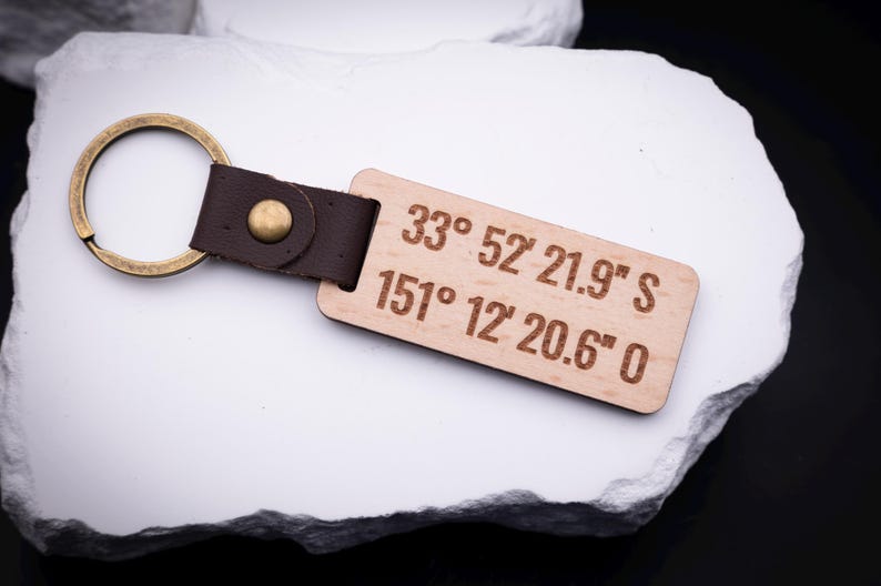 in gravierter Holz Schlüsselanhänger mit den Koordinaten „33° 52' 21.9'' S, 151° 12' 20.6'' O“ liegt auf weißem Stein. Der Anhänger ist aus hellem Holz gefertigt, mit braunem Lederband und stabilem Metallring in Antik-Gold versehen. Perfekt für personalisierte Gravuren von besonderen Orten, Namen oder Sprüchen. Ein liebevolles Geschenk für Partner, Familie oder Freunde, das praktische Funktionalität mit emotionalem Wert verbindet.