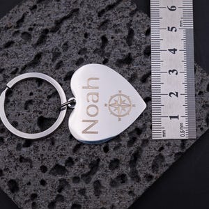 Detailaufnahme des gravierten Herzanhängers aus Edelstahl mit Namensgravur „Noah“ und Kompasssymbol neben Maßlineal. Das Maß zeigt die kompakte Größe des Anhängers, ideal für Schlüsselbund oder Tasche. Robustes Material, präzise Lasergravur und elegantes Design – ein persönliches, langlebiges Geschenk.