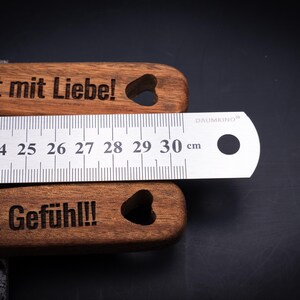 Detailaufnahme des gravierten Akazienholz-Kochlöffelsets mit Edelstahllineal zur Längenmessung. Die Gravuren „Sophia wendet mit Liebe!“ und „Moritz rührt mit Gefühl!!!“ zeigen die hochwertige Lasergravur in edlem Kontrast. Das Maßband zeigt eine Länge von 30 cm – perfekt für personalisierte Küchenutensilien aus echtem Holz.