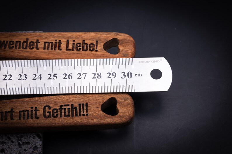 Detailaufnahme des gravierten Akazienholz-Kochlöffelsets mit Edelstahllineal zur Längenmessung. Die Gravuren „Sophia wendet mit Liebe!“ und „Moritz rührt mit Gefühl!!!“ zeigen die hochwertige Lasergravur in edlem Kontrast. Das Maßband zeigt eine Länge von 30 cm – perfekt für personalisierte Küchenutensilien aus echtem Holz.