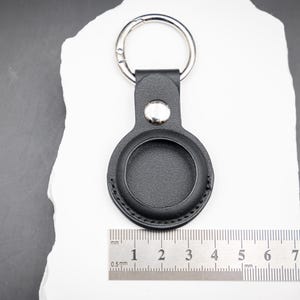 Rückseite eines schwarzen AirTag Schlüsselanhängers aus hochwertigem PU Leder, flach liegend auf weißem Stein mit horizontalem Lineal zur Darstellung der exakten Maße. Die runde AirTag Halterung ist sauber eingefasst und zeigt die glatte Rückseite ohne Gravur. Der robuste Metallring und der Druckknopf sind gut erkennbar. Das Foto vermittelt Größe, Materialstärke und Verarbeitung und eignet sich zur realistischen Einschätzung der Abmessungen vor dem Kauf.