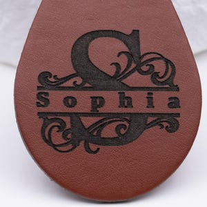 Detailaufnahme eines personalisierten Schlüsselanhängers aus braunem Kunstleder mit feiner Lasergravur. Das kunstvolle Monogramm mit großem Anfangsbuchstaben und geschwungenen Ornamenten zeigt den Namen „Sophia“ in elegantem Design. Die Gravur wirkt präzise und kontrastreich auf der glatten Lederoberfläche – ideal als Geschenkidee für Geburtstage, Jubiläen oder zur stilvollen Personalisierung des eigenen Schlüsselbunds.