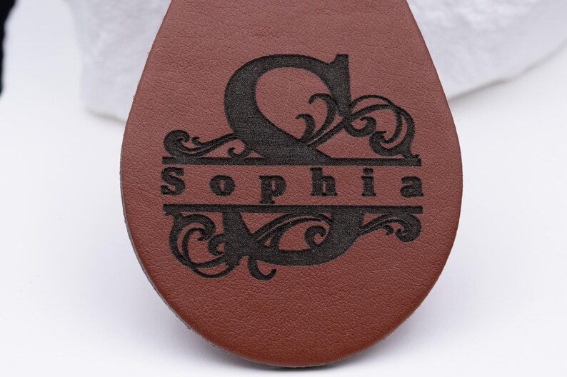 Detailaufnahme eines personalisierten Schlüsselanhängers aus braunem Kunstleder mit feiner Lasergravur. Das kunstvolle Monogramm mit großem Anfangsbuchstaben und geschwungenen Ornamenten zeigt den Namen „Sophia“ in elegantem Design. Die Gravur wirkt präzise und kontrastreich auf der glatten Lederoberfläche – ideal als Geschenkidee für Geburtstage, Jubiläen oder zur stilvollen Personalisierung des eigenen Schlüsselbunds.