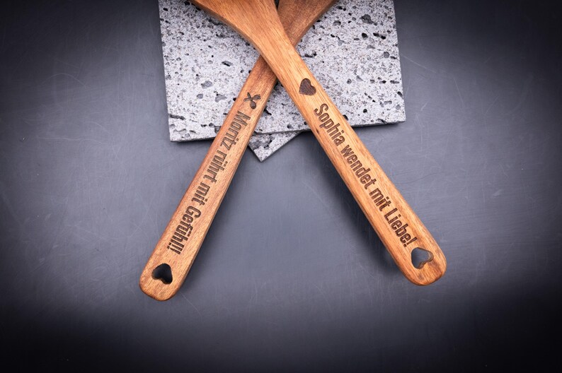 Kochlöffelset aus Akazienholz mit Gravur, bestehend aus einem Pfannenwender und einem Löffel. Beide sind personalisiert mit Namen und Spruch. Das natürliche Holz, die feine Gravur und das Herzloch im Griff machen das Set zu einem beliebten, praktischen Geschenk für Paare und Küchenliebhaber.