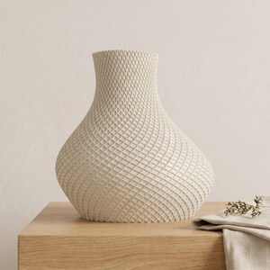 May include: A cream-colored vase with a textured, woven pattern. The vase has a rounded body and a narrow neck. It sits on a light wood surface, with a folded linen cloth and dried sprig of greenery nearby. The vase is a decorative home accent.