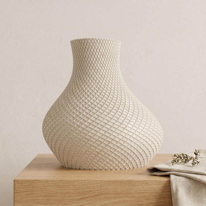 May include: A cream-colored vase with a textured, woven pattern. The vase has a rounded body and a narrow neck. It sits on a light wood surface, with a folded linen cloth and dried sprig of greenery nearby. The vase is a decorative home accent.