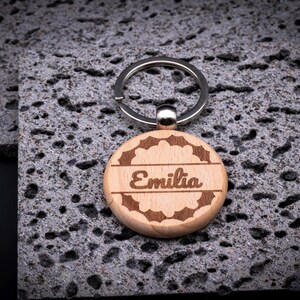 Runder Schlüsselanhänger aus Holz mit Gravur „Emilia“. Verspielter Rahmen mit floralen Ornamenten umgibt den Namen. Edles Design, stabiler Metallring, ideal als individuelles Geschenk.