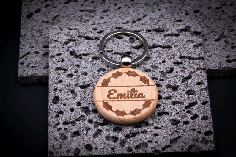 Runder Schlüsselanhänger aus Holz mit Gravur „Emilia“. Verspielter Rahmen mit floralen Ornamenten umgibt den Namen. Edles Design, stabiler Metallring, ideal als individuelles Geschenk.