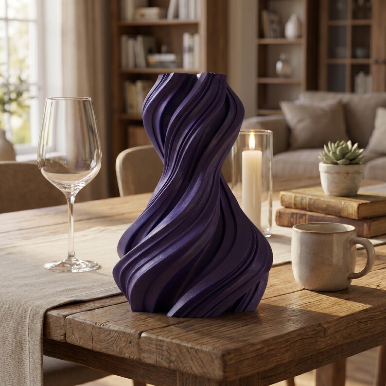 K&ouml;nnte beinhalten: Eine dunkelviolette, skulpturale Vase mit einem gedrehten Design steht auf einem Holztisch. Die Vase hat eine strukturierte, geriffelte Oberfl&auml;che. Ein Weinglas, eine Tasse und eine Kerze stehen ebenfalls auf dem Tisch, was auf ein Wohndekor-Thema hindeutet.