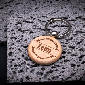 Runder Schlüsselanhänger aus Holz mit Gravur „Leon“. Das Motiv zeigt einen dekorativen Rahmen im rustikalen Stil, mittig der Name. Holzanhänger mit Metallring – natürlich, stabil und personalisierbar.