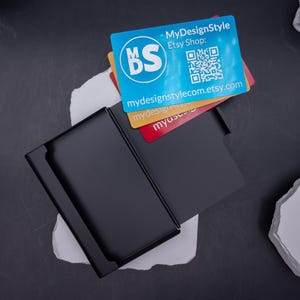 Geöffnetes, leeres Visitenkartenetui aus schwarzem Metall auf weißem Stein fotografiert. Schlichtes, minimalistisches Design, robust und leicht. Ideal für die Aufbewahrung von Visitenkarten, Bankkarten oder Kreditkarten. Kann individuell mit Lasergravur personalisiert werden. Perfekt für Geschäftsleute, die ein stilvolles Accessoire im Büro oder bei Meetings nutzen möchten.