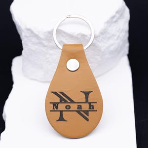 Personalisierter Schlüsselanhänger aus hellbraunem veganem Kunstleder mit Gravur des Namens „Noah“ im Monogramm-Stil. Das große Initial „N“ ist kunstvoll gestaltet, die Gravur in Schwarz kontrastiert edel mit dem warmen Lederfarbton. Der Anhänger liegt auf einem weißen Steinuntergrund, der das hochwertige Material und die feine Verarbeitung betont. Perfekt als Geschenk für Männer oder Frauen.