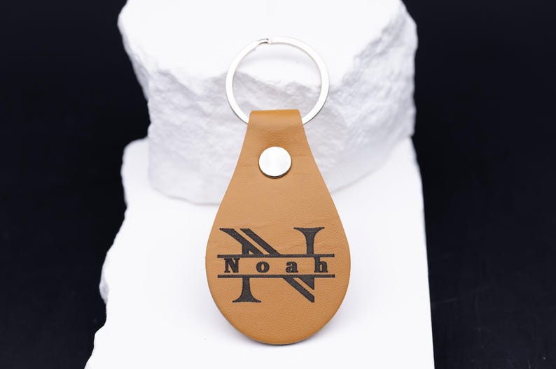 Personalisierter Schlüsselanhänger aus hellbraunem veganem Kunstleder mit Gravur des Namens „Noah“ im Monogramm-Stil. Das große Initial „N“ ist kunstvoll gestaltet, die Gravur in Schwarz kontrastiert edel mit dem warmen Lederfarbton. Der Anhänger liegt auf einem weißen Steinuntergrund, der das hochwertige Material und die feine Verarbeitung betont. Perfekt als Geschenk für Männer oder Frauen.