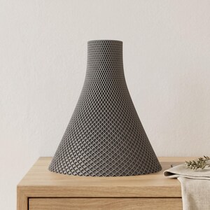 Peut inclure: Un vase gris en forme de c&ocirc;ne avec un motif en treillis. Le vase a une base large qui se r&eacute;tr&eacute;cit vers le haut. Il est fait d'un mat&eacute;riau &agrave; l'aspect textur&eacute; et repose sur une surface en bois.
