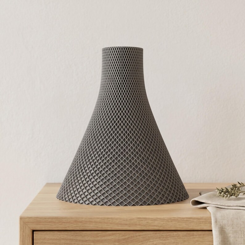 Peut inclure: Un vase gris en forme de c&ocirc;ne avec un motif en treillis. Le vase a une base large qui se r&eacute;tr&eacute;cit vers le haut. Il est fait d'un mat&eacute;riau &agrave; l'aspect textur&eacute; et repose sur une surface en bois.