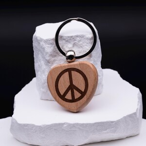 Herz Schlüsselanhänger aus Holz mit eingraviertem Peace-Symbol. Die feine Gravur verleiht dem Anhänger eine zeitlose und moderne Optik. Personalisierbar mit weiteren Namen oder Symbolen eignet er sich als Geschenk für Freunde, Paare oder Kinder. Ein nachhaltiges Accessoire, das durch schlichte Eleganz und Individualität überzeugt und sowohl im Alltag als auch als besonderes Geschenk Freude bereitet.
