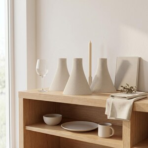 Peut inclure: Trois vases d&eacute;coratifs beiges en forme de c&ocirc;ne avec une surface textur&eacute;e sont expos&eacute;s sur une &eacute;tag&egrave;re en bois. Un verre &agrave; vin, une bougie, un petit bol, une assiette, une tasse et une serviette en lin pli&eacute;e compl&egrave;tent le d&eacute;cor minimaliste.