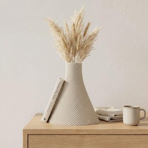 Peut inclure: Un vase beige texturé avec un motif géométrique contient des herbes de la pampa séchées. Un livre, des serviettes pliées et une tasse sont posés sur une surface en bois. Le vase a une base large et un col étroit.