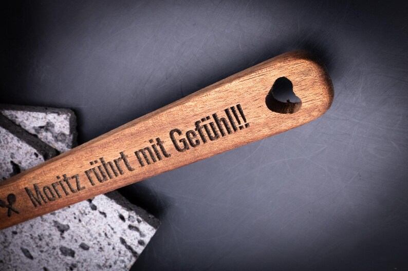 Detailaufnahme des gravierten Holzlöffels aus Akazienholz mit der Gravur „Moritz rührt mit Gefühl!!!“ und kleinem Symbol. Das Herzloch im Griff sorgt für dekorative Aufhängemöglichkeit. Die feine Lasergravur ist dauerhaft eingebrannt – ideal für personalisierte Geschenke zum Kochen, Braten oder Backen.