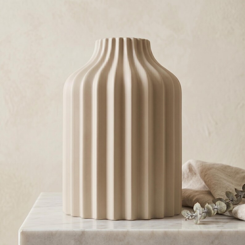 Może przedstawiać: Beżowy ceramiczny wazon z pionowymi żebrowanymi detalami. Wazon ma zaokrąglony korpus i lekko rozszerzającą się szyję. Jest prezentowany na białej marmurowej powierzchni. Dekoracyjny element wystroju domu.