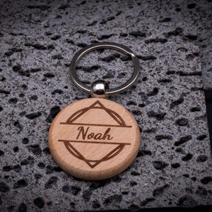 Runder Schlüsselanhänger aus Holz mit Gravur „Noah“. Moderner geometrischer Rahmen mit klaren Linien. Robuster Anhänger mit Metallring, perfekt als personalisierter Begleiter.