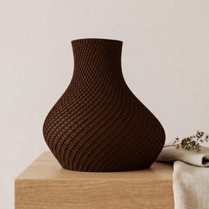 May include: A brown decorative vase with a lattice design. The vase has a wide base that tapers to a narrow neck. The vase is sitting on a light brown wooden surface. A beige cloth and sprig of greenery are in the background.