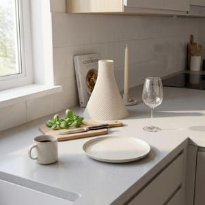 Peut inclure: Sc&egrave;ne de cuisine avec un livre de cuisine, un vase d&eacute;coratif blanc textur&eacute;, une planche &agrave; d&eacute;couper avec du basilic et un couteau, une tasse, une assiette, un verre &agrave; vin et une bougie. Le livre de cuisine est ouvert sur une page avec une photo de nourriture.