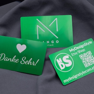 Drei hochwertige Visitenkarten aus grün eloxiertem Aluminium liegen auf grauem Stoff. Die Karten zeigen verschiedene Designs: eine mit Herz und "Danke Sehr!", eine mit Firmenlogo und eine mit QR-Code für den Etsy-Shop. Die stabile Metalloberfläche sorgt für edlen Glanz und verleiht den Karten ein modernes, professionelles Auftreten, ideal als langlebige Geschäftsausstattung.