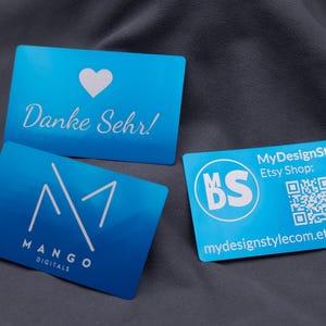 Drei Visitenkarten aus Aluminium in elegantem Blau: eine Dankeskarte mit Herzsymbol und Gravur „Danke Sehr!“, eine Firmenkarte von Mango Digitals mit Logo sowie eine MyDesignStyle-Karte mit QR-Code und Etsy-Shop-Adresse. Die matten Metallkarten mit abgerundeten Kanten wirken edel, stabil und modern. Ideal als exklusive Visitenkarten, Kundenkarten oder stilvolle Dankeskarten für Unternehmen und kreative Shops.