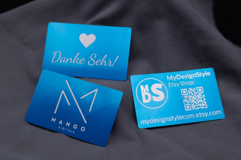 Drei Visitenkarten aus Aluminium in elegantem Blau: eine Dankeskarte mit Herzsymbol und Gravur „Danke Sehr!“, eine Firmenkarte von Mango Digitals mit Logo sowie eine MyDesignStyle-Karte mit QR-Code und Etsy-Shop-Adresse. Die matten Metallkarten mit abgerundeten Kanten wirken edel, stabil und modern. Ideal als exklusive Visitenkarten, Kundenkarten oder stilvolle Dankeskarten für Unternehmen und kreative Shops.
