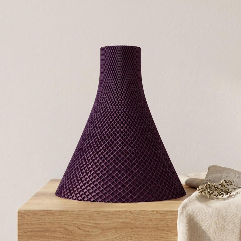 Peut inclure: Un vase d&eacute;coratif violet fonc&eacute; avec un motif g&eacute;om&eacute;trique en treillis. Le vase a une base conique large qui se r&eacute;tr&eacute;cit en un col &eacute;troit. Il est pos&eacute; sur une surface en bois de couleur claire, avec un fond neutre.
