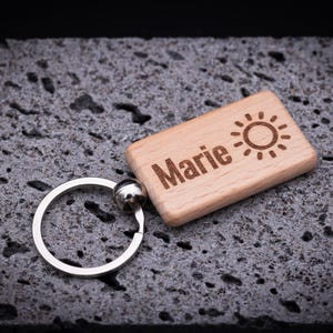Personaliserter Holz-Schlüsselanhänger mit rechteckiger Form, ca. 5 cm Länge, graviert mit dem Namen Marie und einem Sonnensymbol. Hergestellt aus hochwertiger Buche, mit abgerundeten Ecken und glatter Oberfläche, ausgestattet mit stabilem Schlüsselring aus Edelstahl. Die Lasergravur sorgt für ein langlebiges und detailreiches Design. Perfekt als Geschenk für Freunde oder Familie und als praktisches, personalisiertes Accessoire.