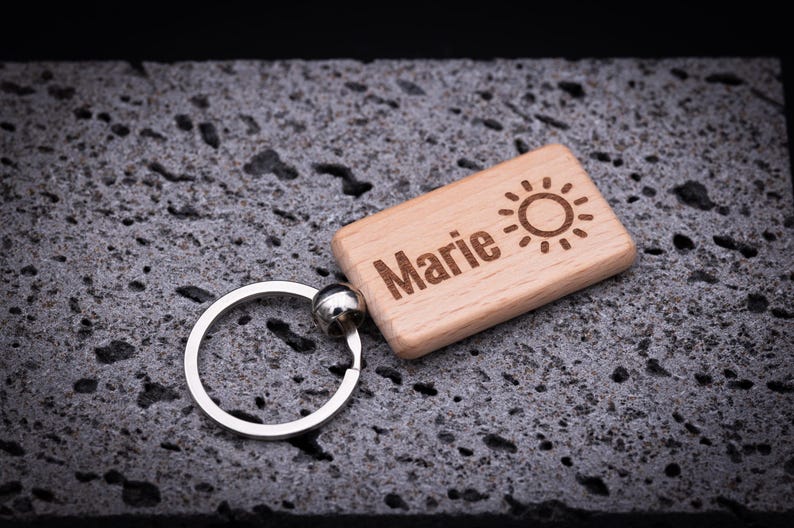Personaliserter Holz-Schlüsselanhänger mit rechteckiger Form, ca. 5 cm Länge, graviert mit dem Namen Marie und einem Sonnensymbol. Hergestellt aus hochwertiger Buche, mit abgerundeten Ecken und glatter Oberfläche, ausgestattet mit stabilem Schlüsselring aus Edelstahl. Die Lasergravur sorgt für ein langlebiges und detailreiches Design. Perfekt als Geschenk für Freunde oder Familie und als praktisches, personalisiertes Accessoire.