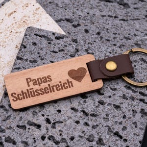 Personalisierter Schlüsselanhänger aus Holz mit Gravur „Papas Schlüsselreich“ und Herz-Symbol. Der rechteckige Anhänger kombiniert natürliche Holzoptik mit braunem Kunstlederband und stabilem Messingring. Ein praktisches und zugleich liebevolles Accessoire für den Schlüsselbund – ideal als Geschenk für Väter zu Geburtstag, Vatertag oder Weihnachten. Langlebig, hochwertig und alltagstauglich.