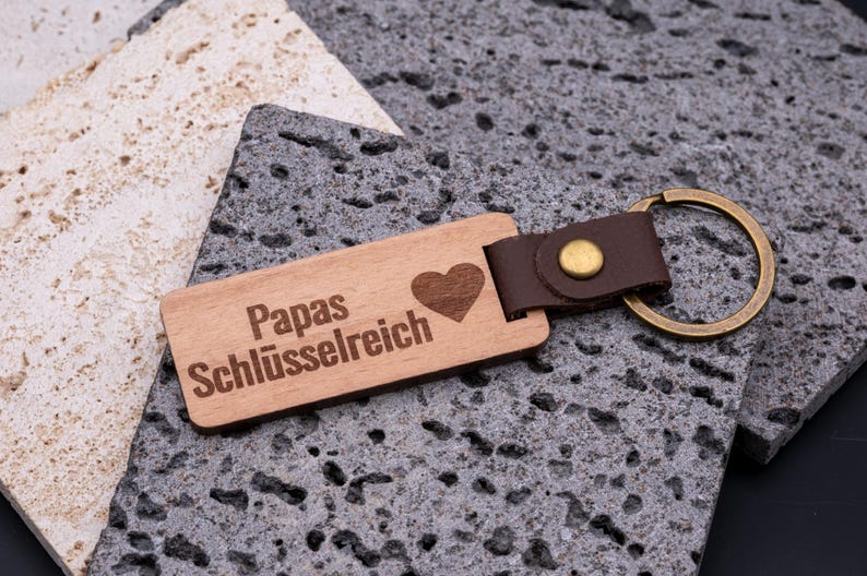 Personalisierter Schlüsselanhänger aus Holz mit Gravur „Papas Schlüsselreich“ und Herz-Symbol. Der rechteckige Anhänger kombiniert natürliche Holzoptik mit braunem Kunstlederband und stabilem Messingring. Ein praktisches und zugleich liebevolles Accessoire für den Schlüsselbund – ideal als Geschenk für Väter zu Geburtstag, Vatertag oder Weihnachten. Langlebig, hochwertig und alltagstauglich.